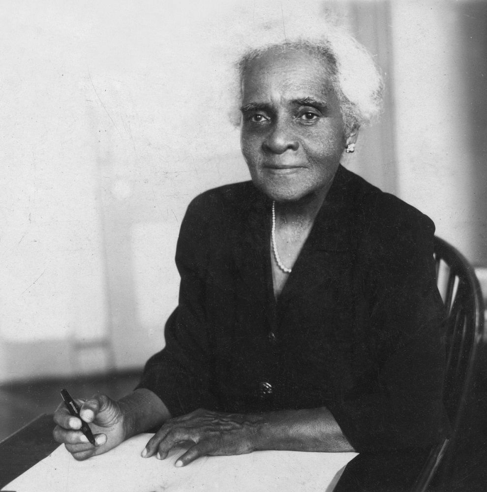 A woman seated and posing with pen to paper